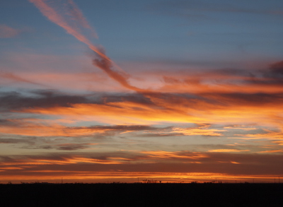 [The sun already gone from view has painted the clouds both at the horizon and further up in the sky shades of yellow, orange, pink, and a yellowish-purple. Light blue sky is visible in the middle of the image and at the top. The ground appears to be a dark orangey-brown.]